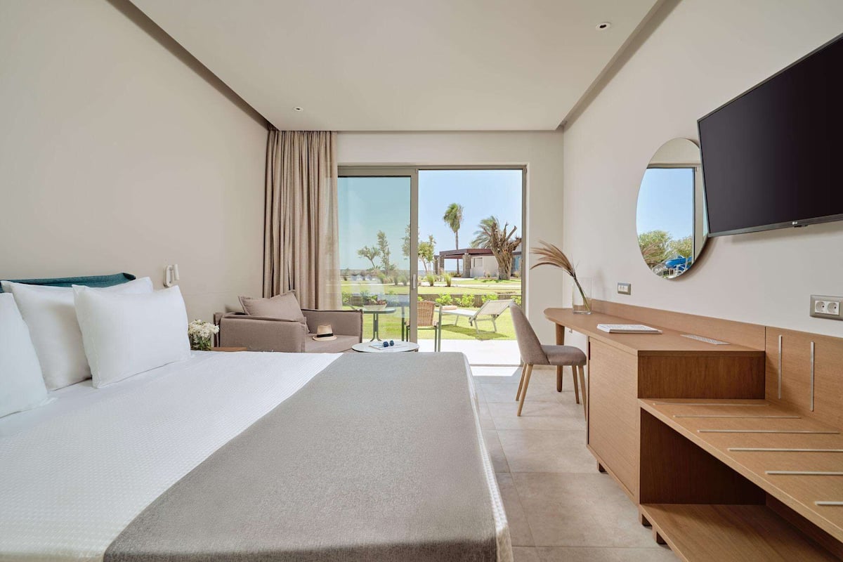 A modern hotel room with a large bed, a desk, a flat-screen TV, and a balcony with a view of the outdoors.