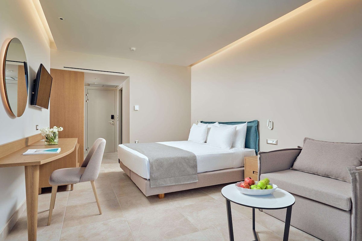 A modern hotel room with a bed, a desk, a chair, a sofa, and a small table with fruit.