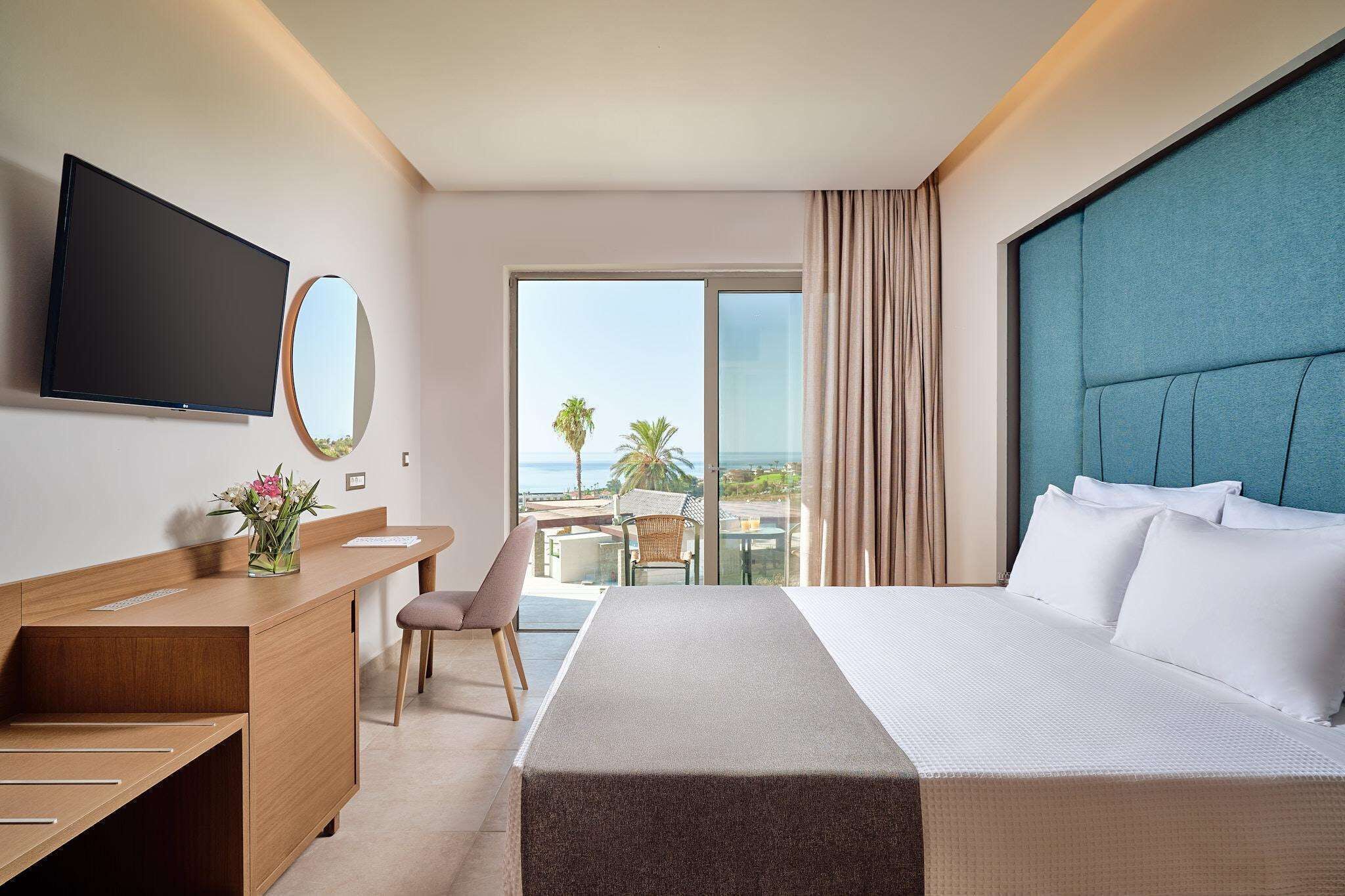 A hotel room with a bed, a desk, a chair, a TV, a mirror, and a view of the ocean.