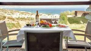 A table set for a meal with a bottle of wine, glasses, and a plate of food, overlooking a scenic view of the sea and hills.