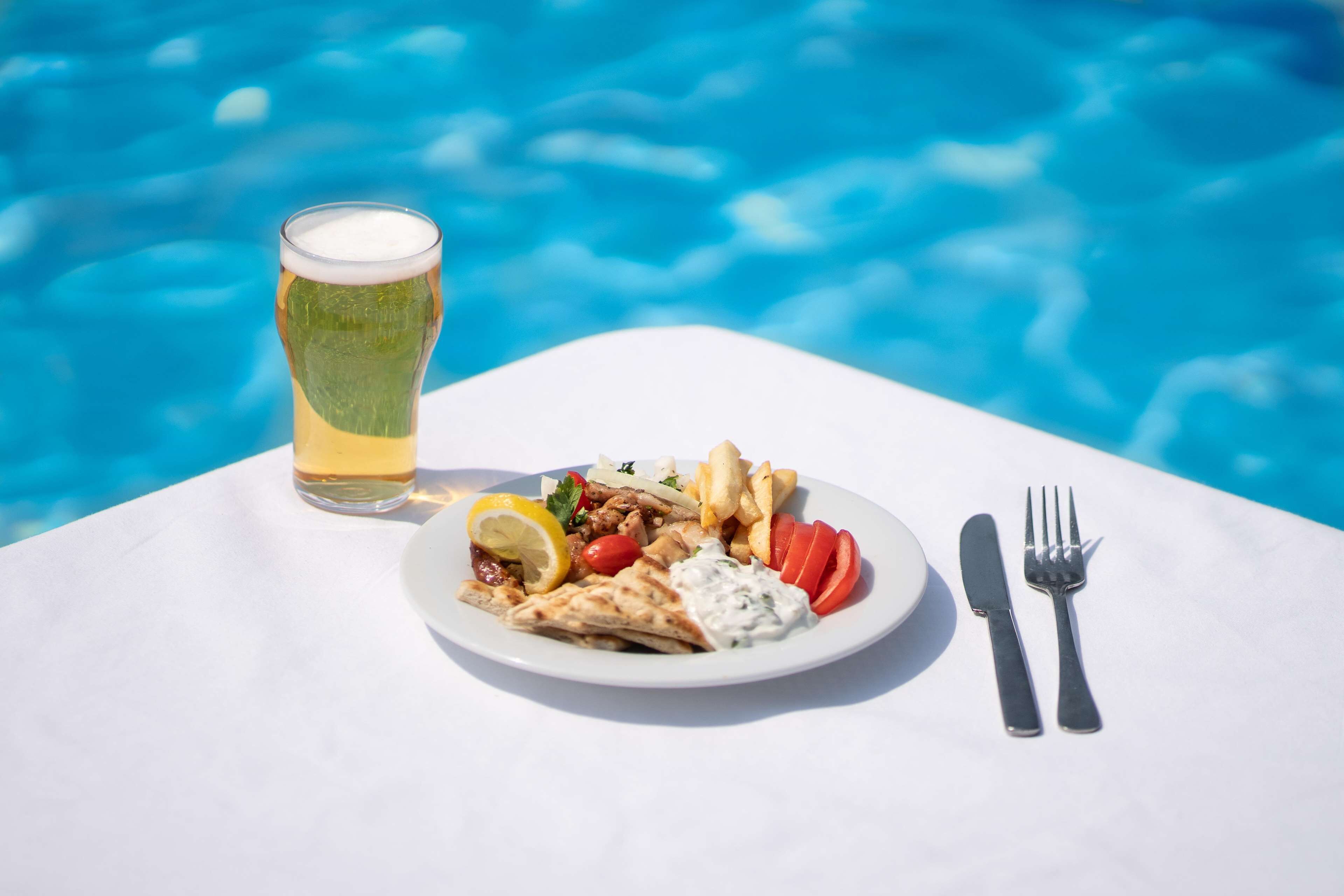 A meal with a glass of beer and a fork and knife on a table by a pool.