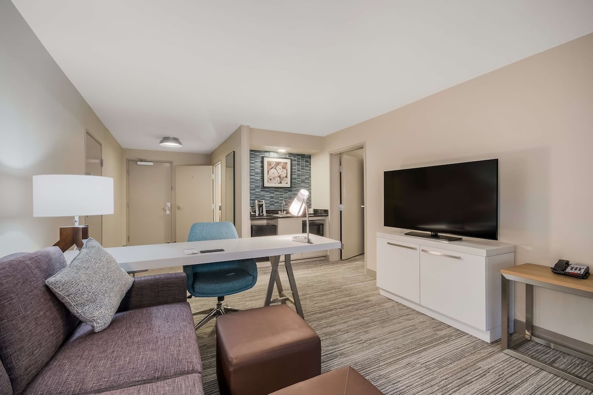 A modern hotel room with a living area, kitchenette, and a flat-screen TV.