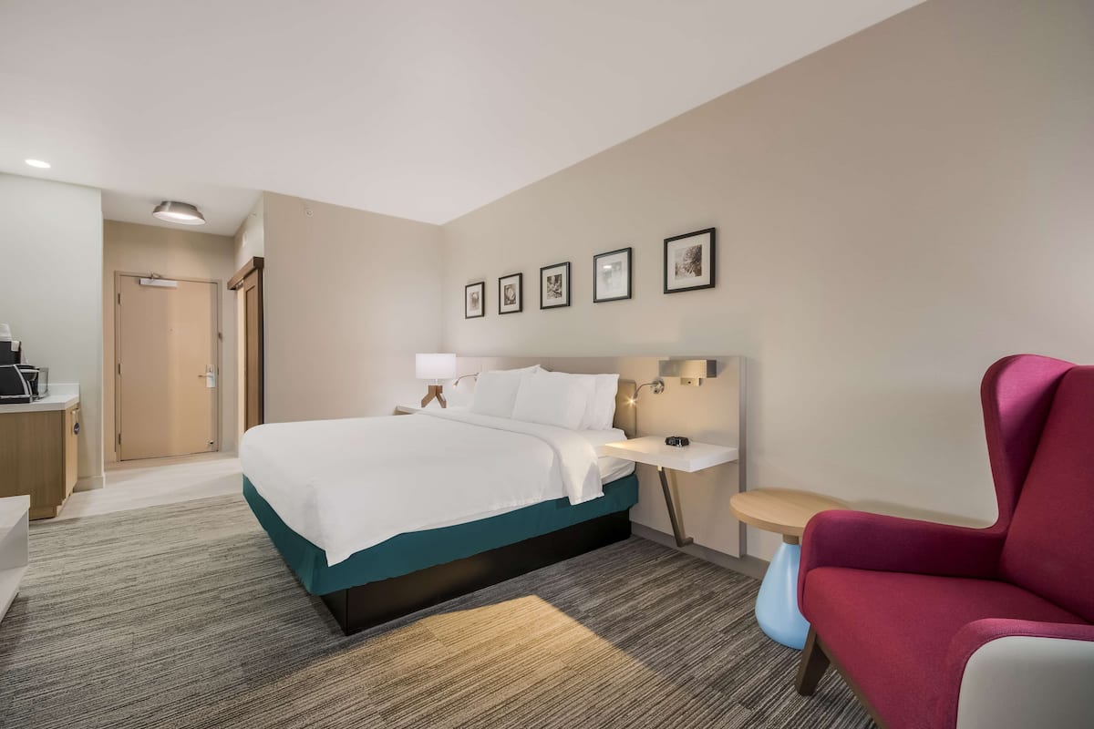 A hotel room with a bed, bedside table, chair, and a desk with a lamp.