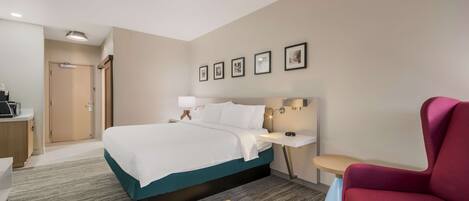 A hotel room with a bed, bedside table, chair, and a desk with a lamp.