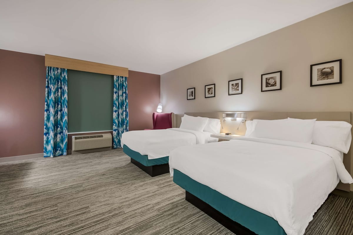 A hotel room with two beds, a nightstand, a lamp, and a wall-mounted heater.