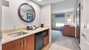 A modern kitchen with a sink, dishwasher, and microwave. There is a large mirror above the sink and a countertop with various items. The room has a neutral color scheme and a view of a living area with a couch and a TV. - Hampton Inn & Suites St. Louis/South I-55 (St. Louis)