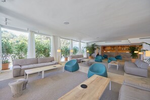 A spacious living area with a sofa, chairs, a coffee table, and a dining table. There are large windows providing natural light and views of greenery outside. - Prinsotel La Caleta Hotel (Ciutadella de Menorca)