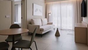 Une chambre d’hôtel moderne équipée d’un canapé, d’une table de salle à manger ronde, d’une petite table d’appoint et d’un téléviseur fixé au mur.