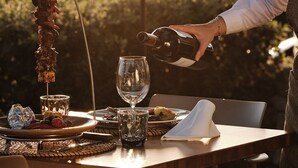 Une table dressée à l’extérieur, avec une bouteille de vin dont on verse le contenu dans un verre, une assiette remplie de nourriture et une brochette décorative.