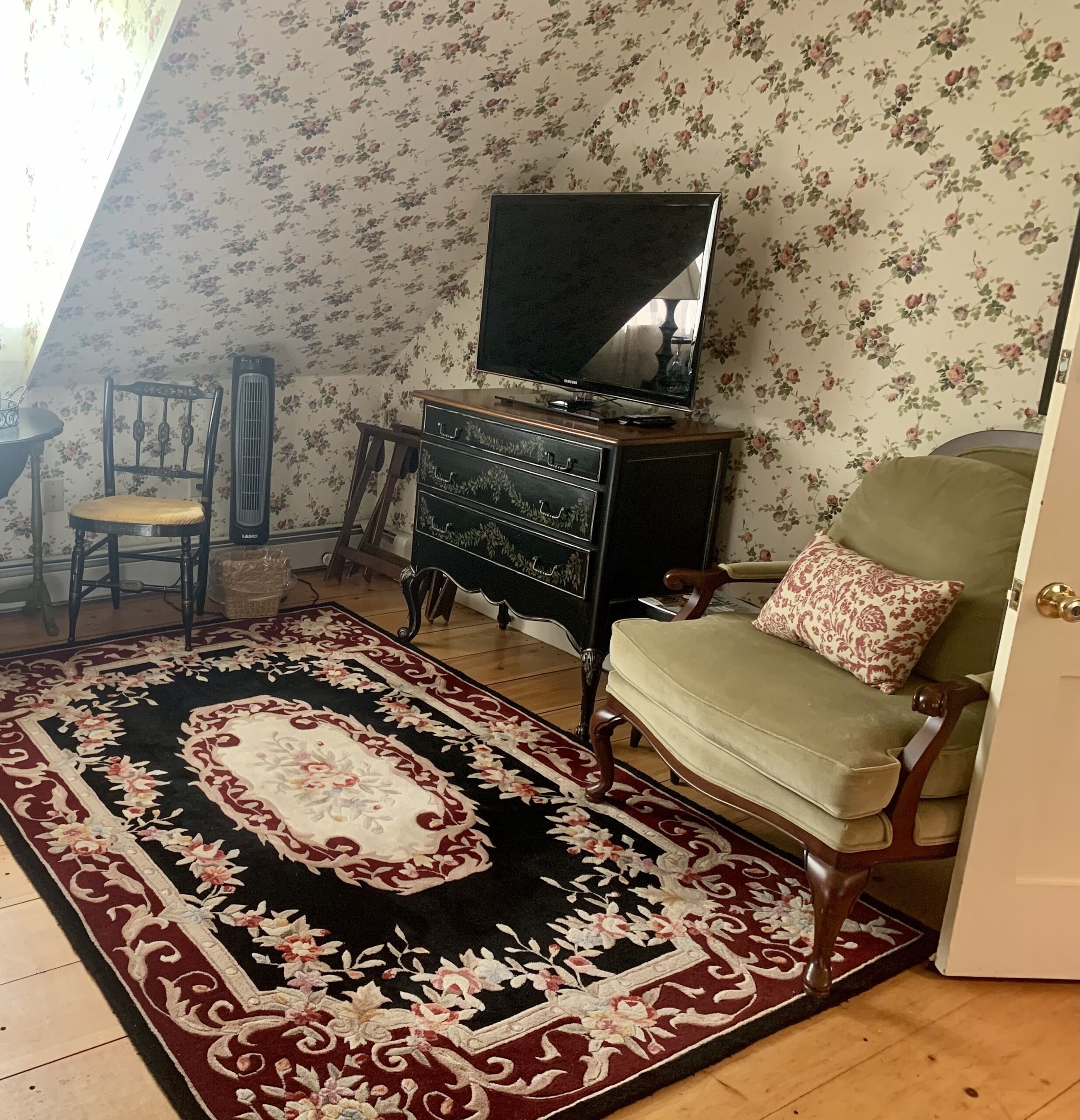 A living room with a floral wallpaper, a television on a wooden stand, a green armchair, and a patterned rug.