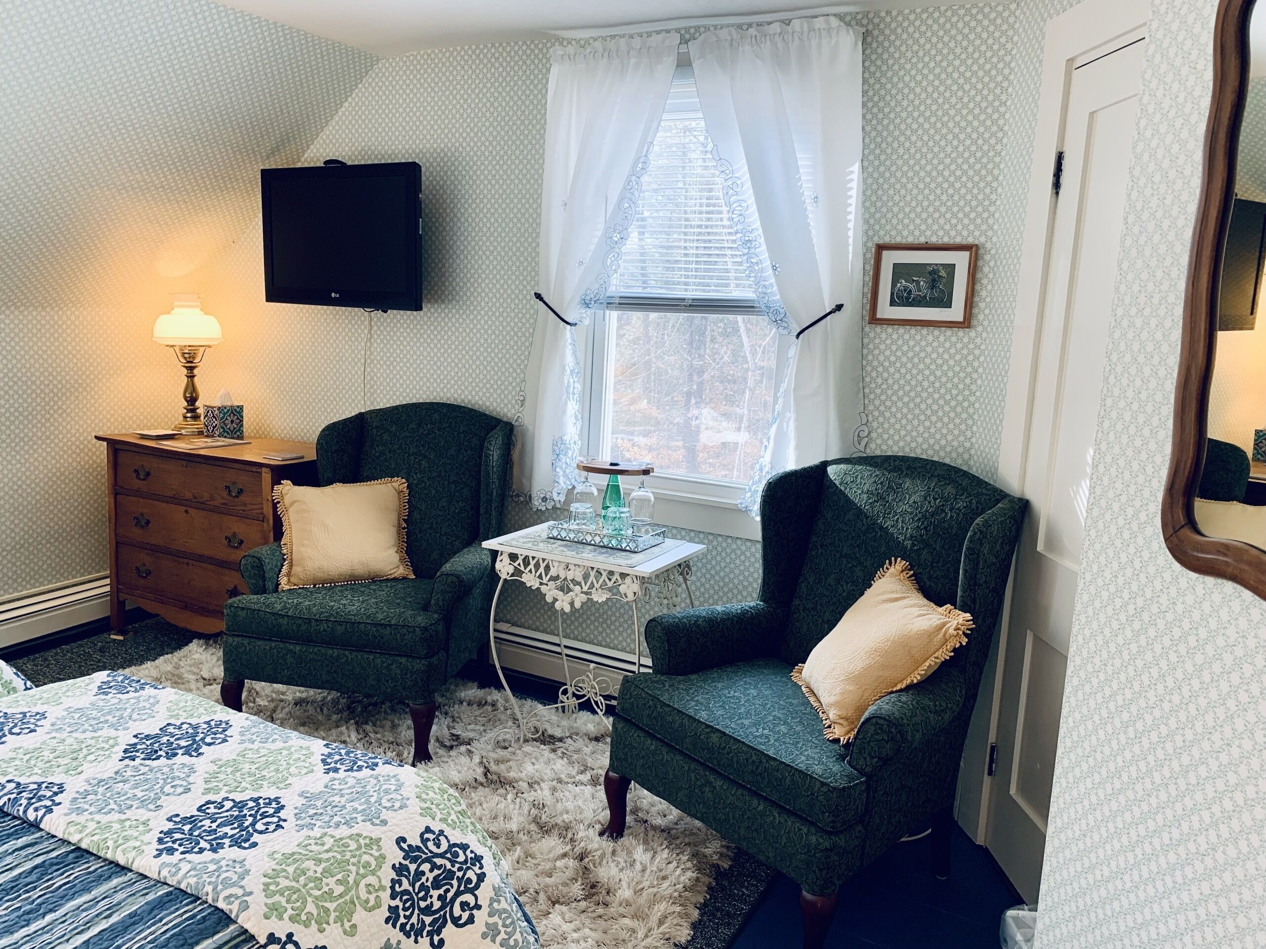 A bedroom with two green armchairs, a wooden dresser, a small table with a lamp, and a TV on the wall.