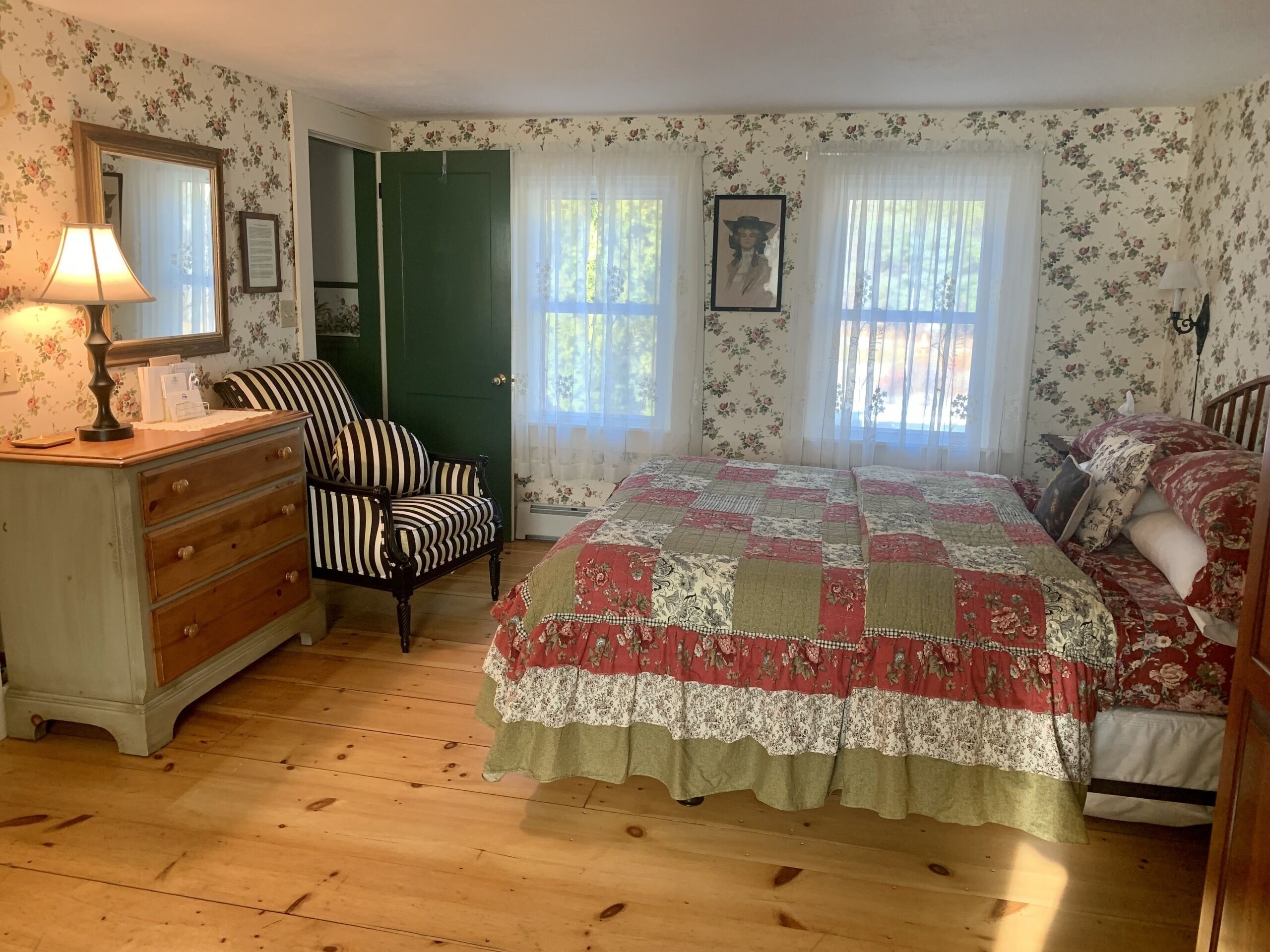 A bedroom with floral wallpaper, a wooden dresser, a striped armchair, a bed with a patchwork quilt, and two windows with sheer curtains.