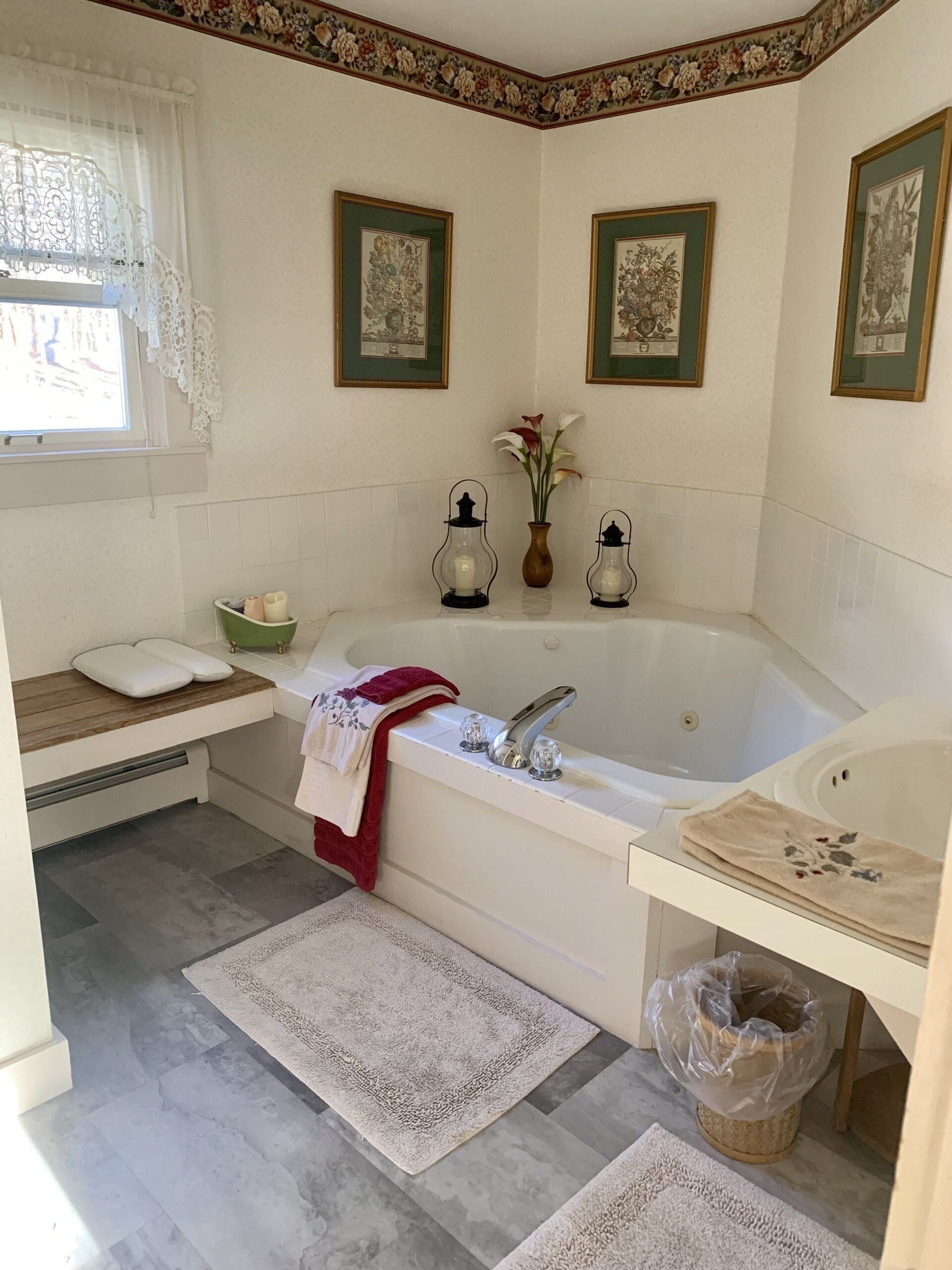 A bathroom with a bathtub, a sink, a window with lace curtains, framed pictures on the wall, and a vase with flowers.