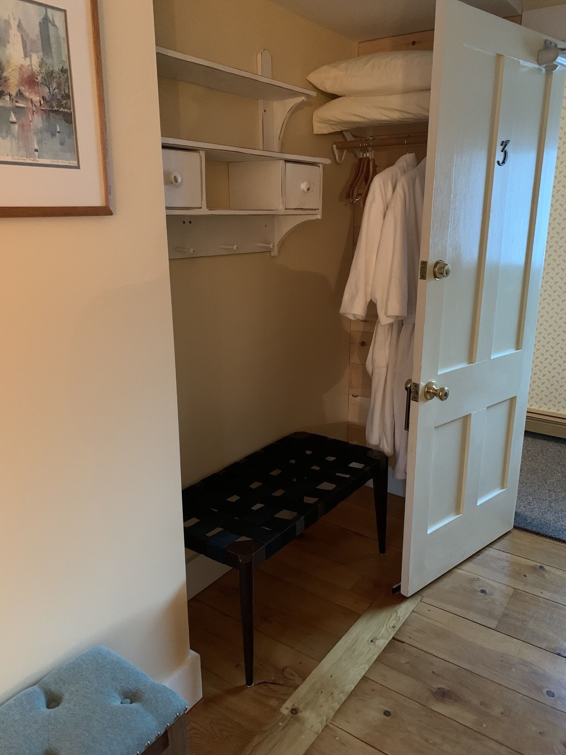 Quarto com um banco de madeira, porta branca com o número 3, cabideiro com roupas penduradas e uma estante com gavetas.