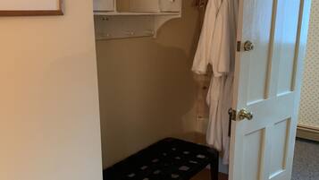 Quarto com um banco de madeira, porta branca com o número 3, cabideiro com roupas penduradas e uma estante com gavetas.