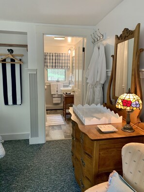 Een slaapkamer met een houten dressoir, een spiegel, een lamp en een stoel. Er is een raam met gordijnen en een handdoekhouder.