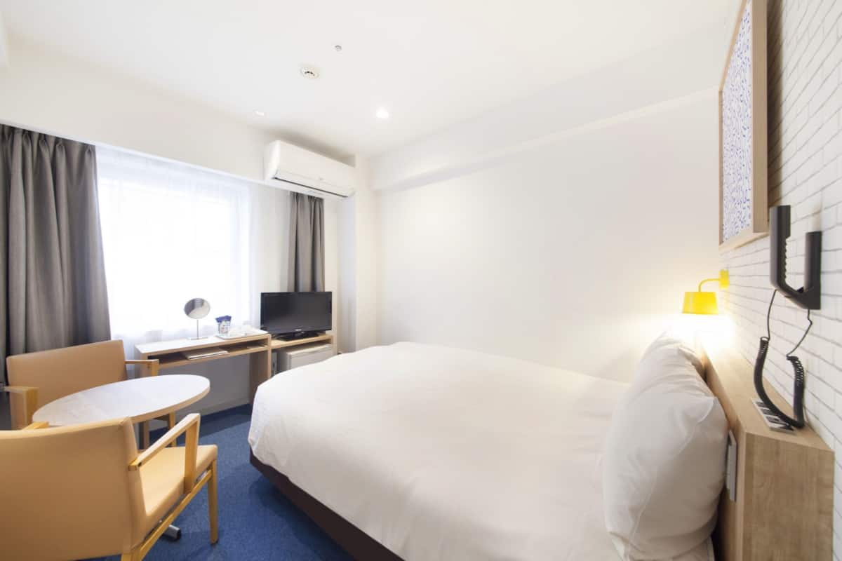 A hotel room with a bed, a desk with a chair, a TV, a lamp, and a phone on the wall.