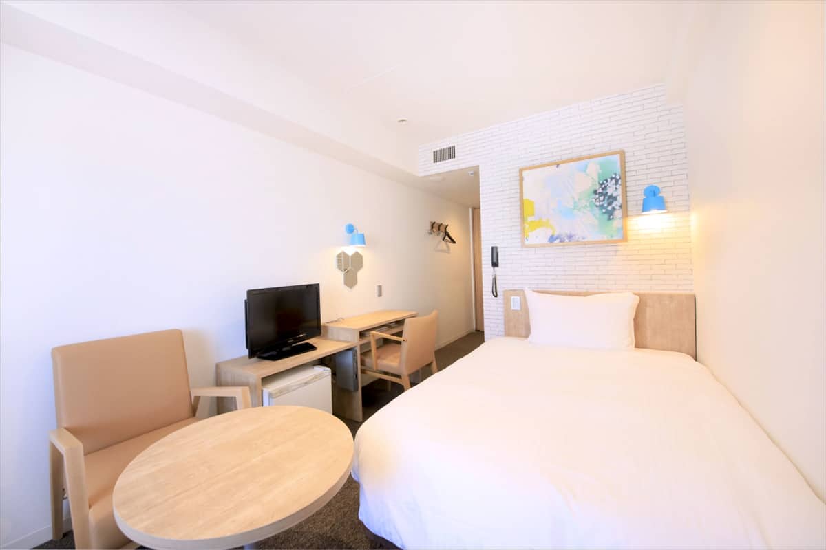 A hotel room with a bed, a desk with a computer, a chair, a small table, and a TV.