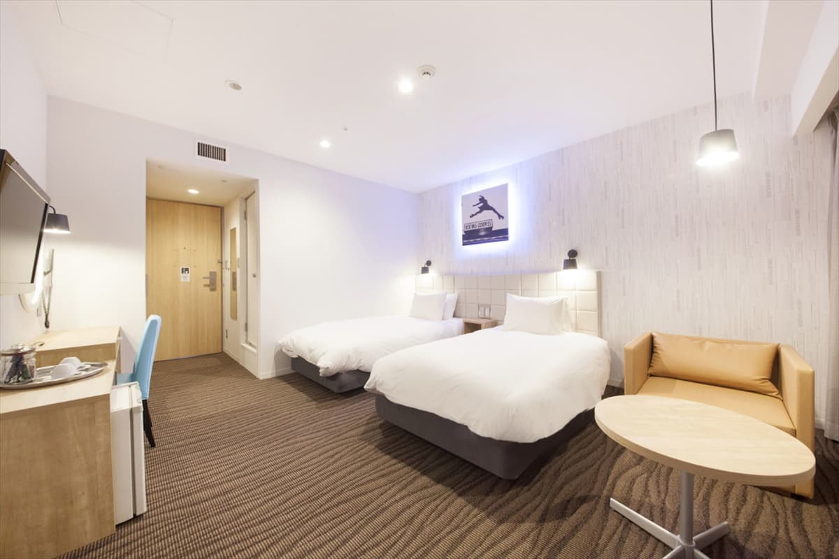 A hotel room with two beds, a desk, a chair, a round table, and a wall-mounted TV.