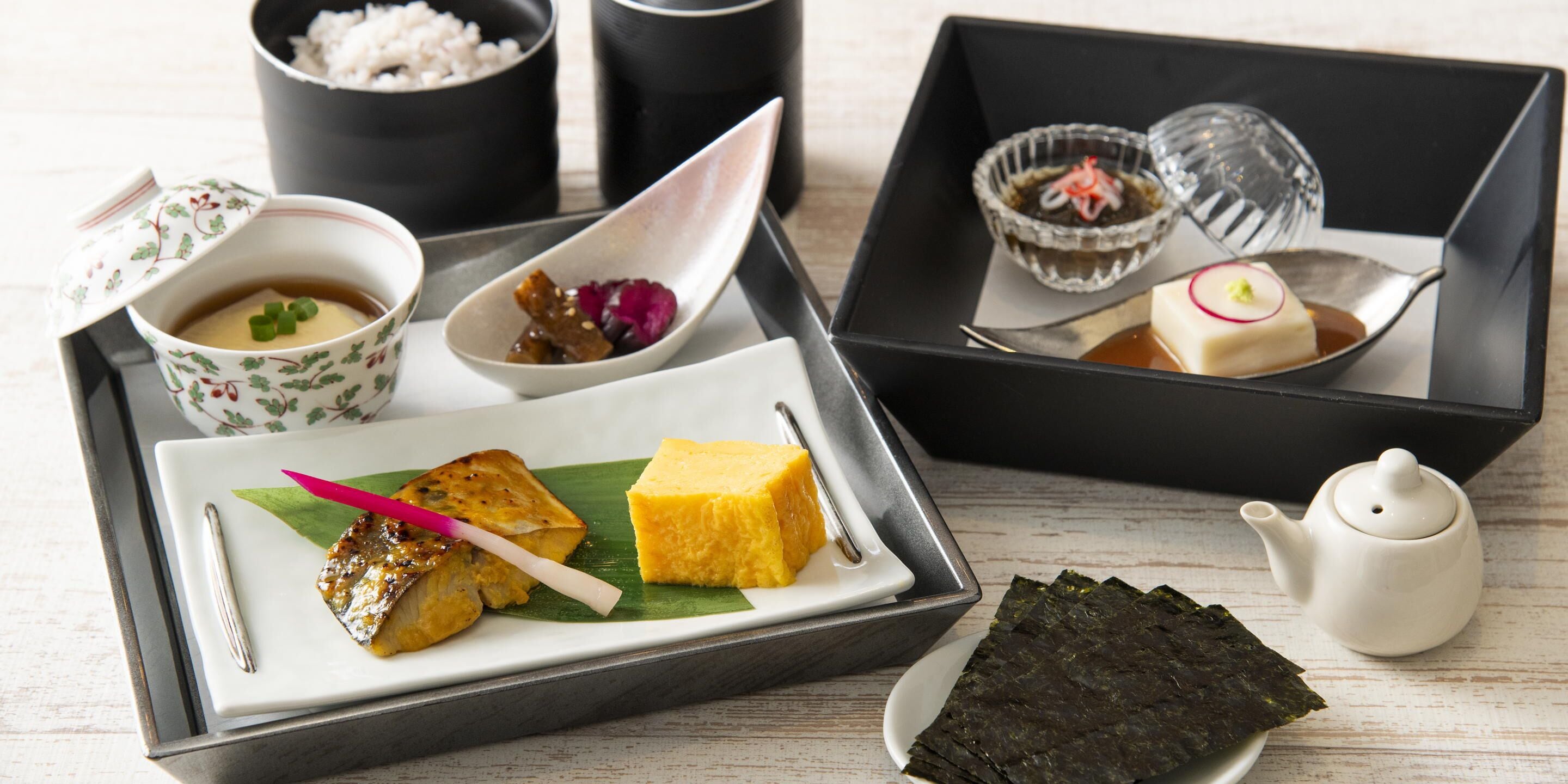 A tray with various Japanese dishes including fish, tofu, and seaweed, accompanied by small bowls of condiments and a teapot.