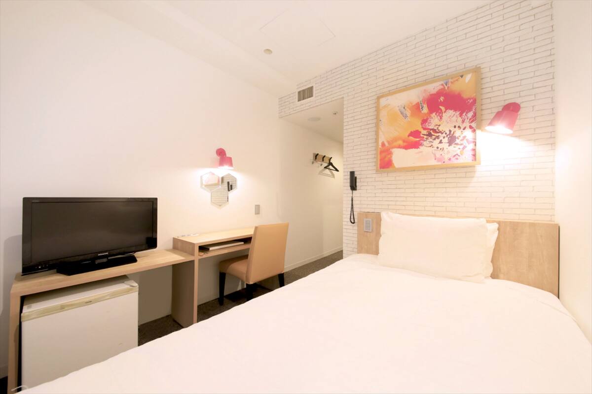 A hotel room with a bed, a desk with a television, a chair, a small refrigerator, and a wall-mounted light.