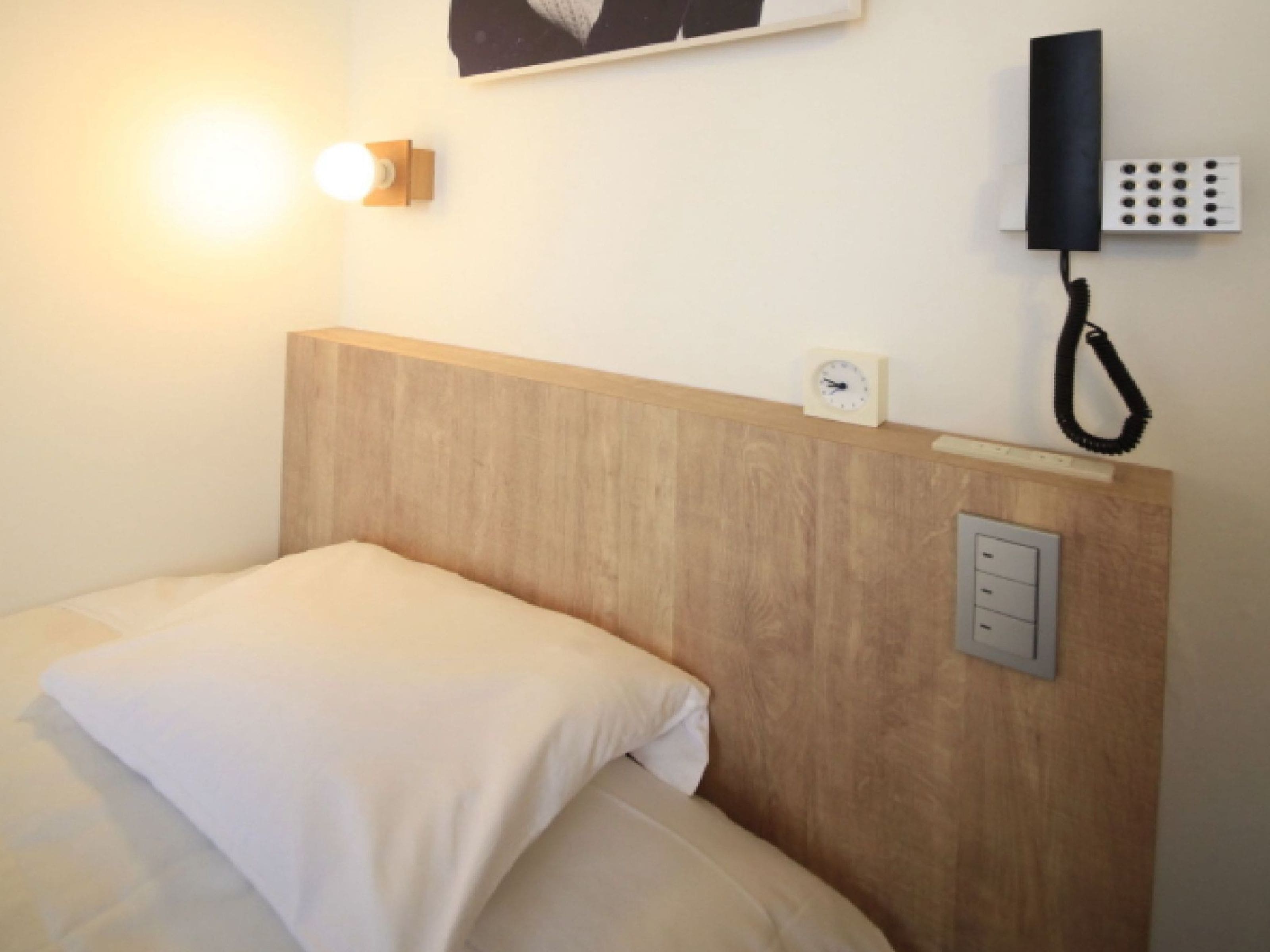 A hotel room with a wooden headboard, a bedside lamp, a telephone, and a clock on the wall.