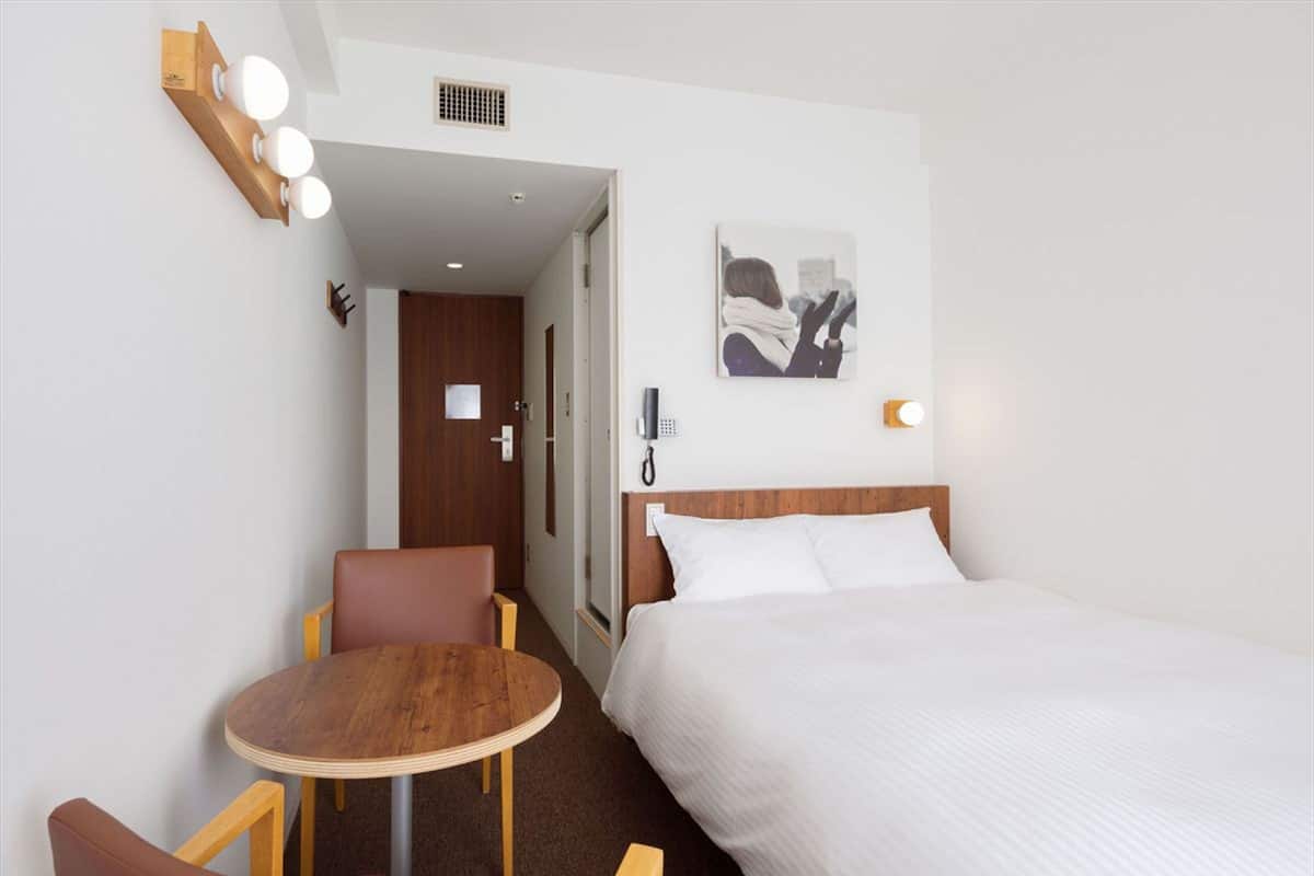 A hotel room with a bed, a desk, and a chair. There is a painting on the wall and a phone on the desk.