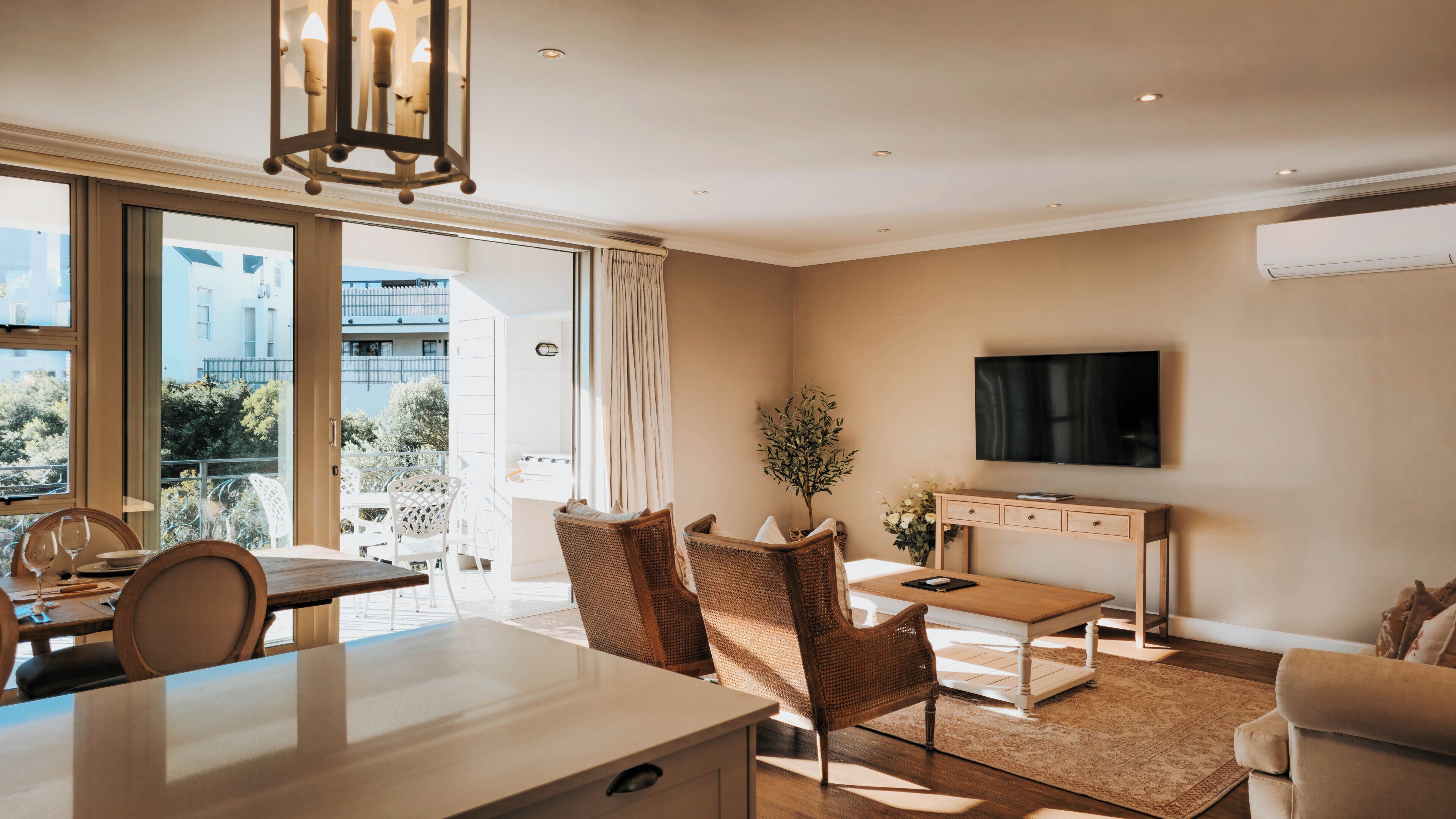 A modern living room with a dining area, large windows, a flat-screen TV, and a balcony.