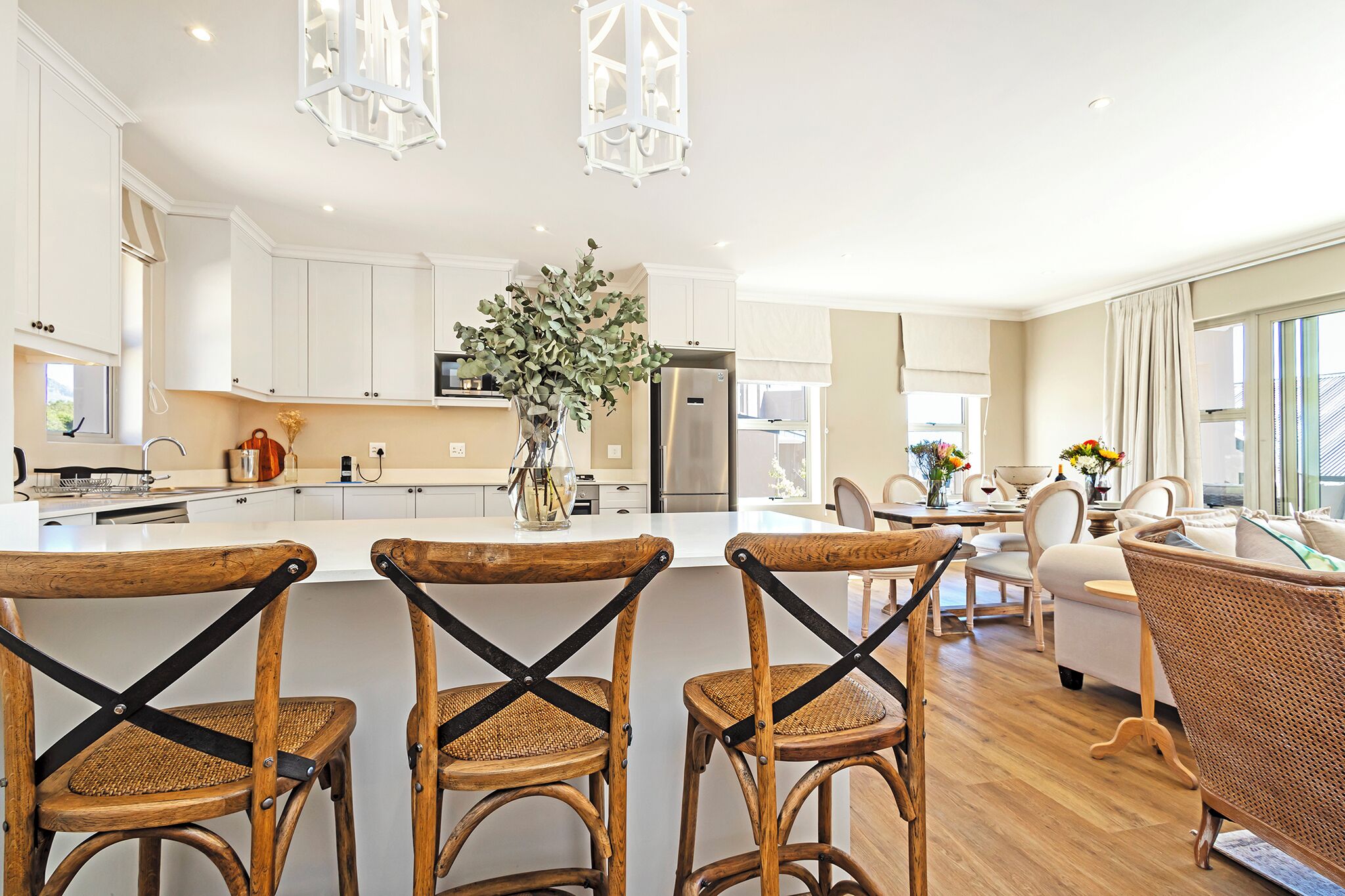 A modern kitchen with white cabinets, a central island with chairs, and a dining area with wicker furniture.