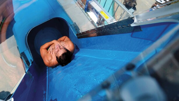 Um toboágua com uma pessoa na base, área de piscina com espreguiçadeiras e um pequeno edifício com telhado verde.