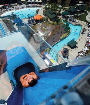 A water slide with a person at the bottom, a pool area with lounge chairs, and a small building with a green roof.
