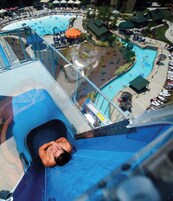 A water slide with a person at the bottom, a pool area with lounge chairs, and a small building with a green roof.