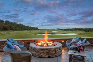 A stone fire pit surrounded by wicker furniture with cushions, a table with glasses and a bottle, and a view of a golf course.