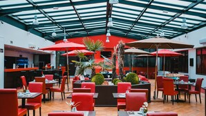 A restaurant with red chairs and tables, a central planter with plants, and a glass ceiling.