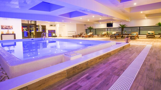 A modern indoor swimming pool with a tiled deck, lounge chairs, and a ceiling with skylights.