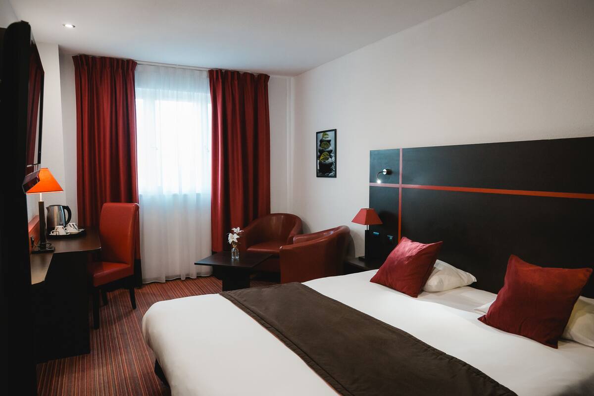 A hotel room with a large bed, red and white pillows, a desk with a lamp, a chair, and a nightstand.