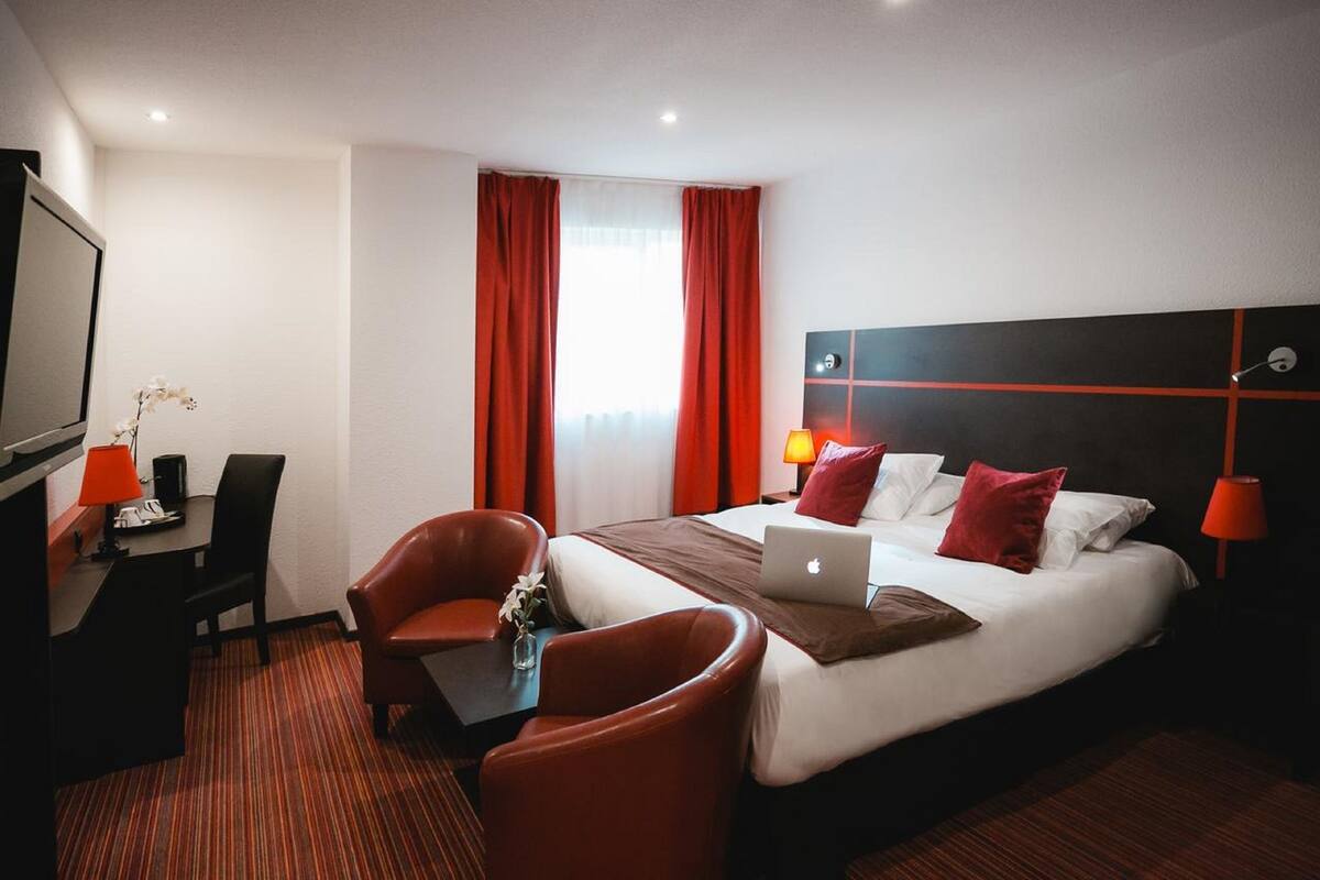 A hotel room with a bed, a desk, a chair, a laptop, and a TV.