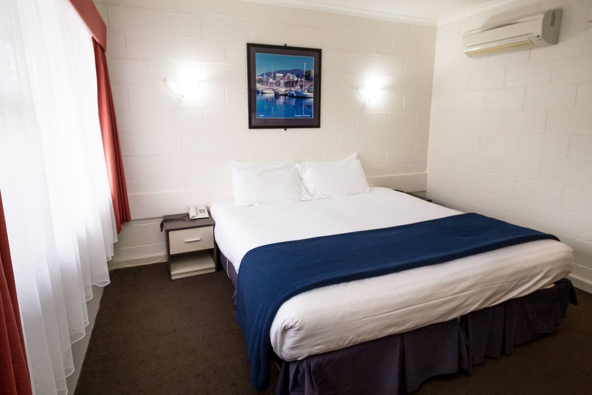 A hotel room with a bed, a nightstand, a telephone, a framed picture on the wall, and an air conditioning unit.