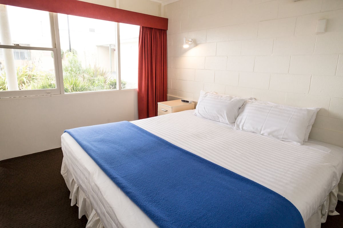 A hotel room with a large bed, a nightstand, a window with curtains, and a brick wall.
