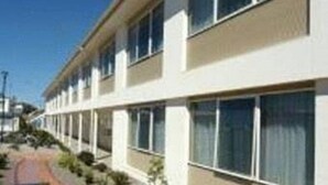 Un edificio moderno con una fachada larga y recta, múltiples ventanas y un paseo pavimentado con plantas pequeñas.