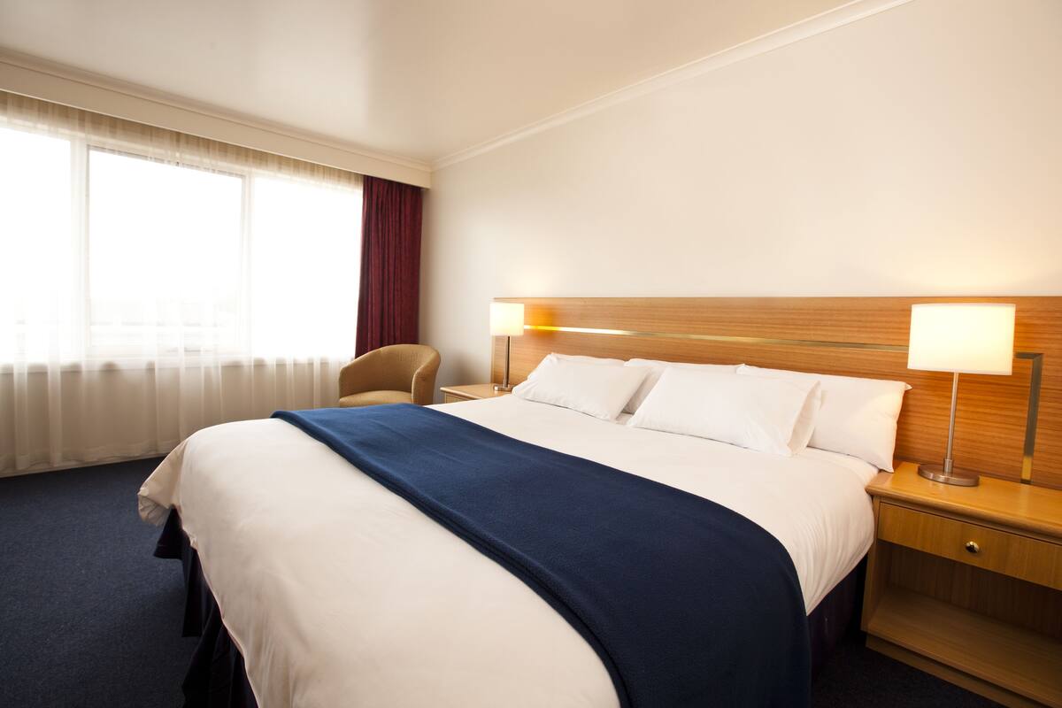 A hotel room with a large bed, two bedside lamps, a wooden nightstand, a chair, and a window with curtains.