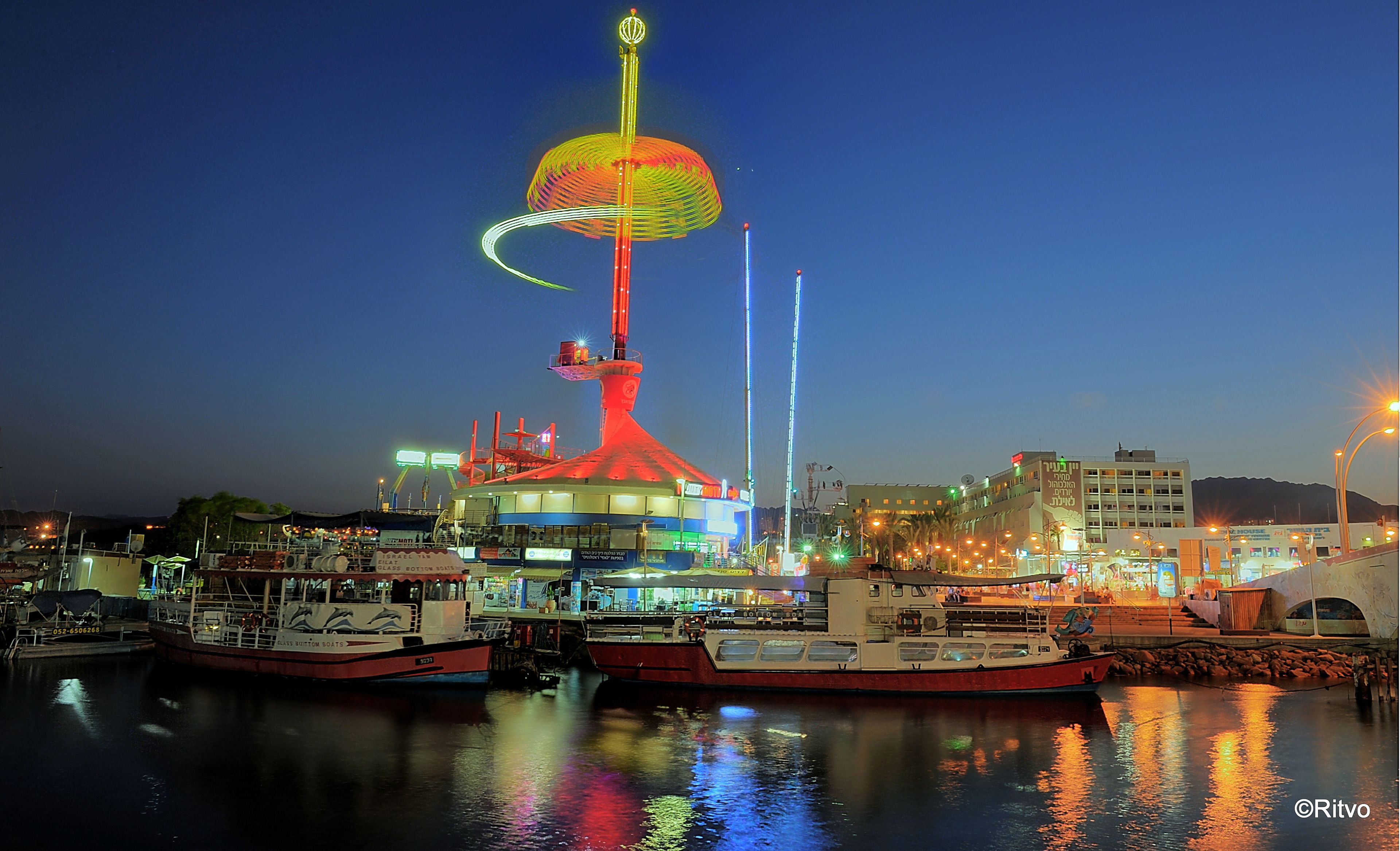A waterfront area with a prominent tower featuring a colorful top, boats docked nearby, and buildings in the background.