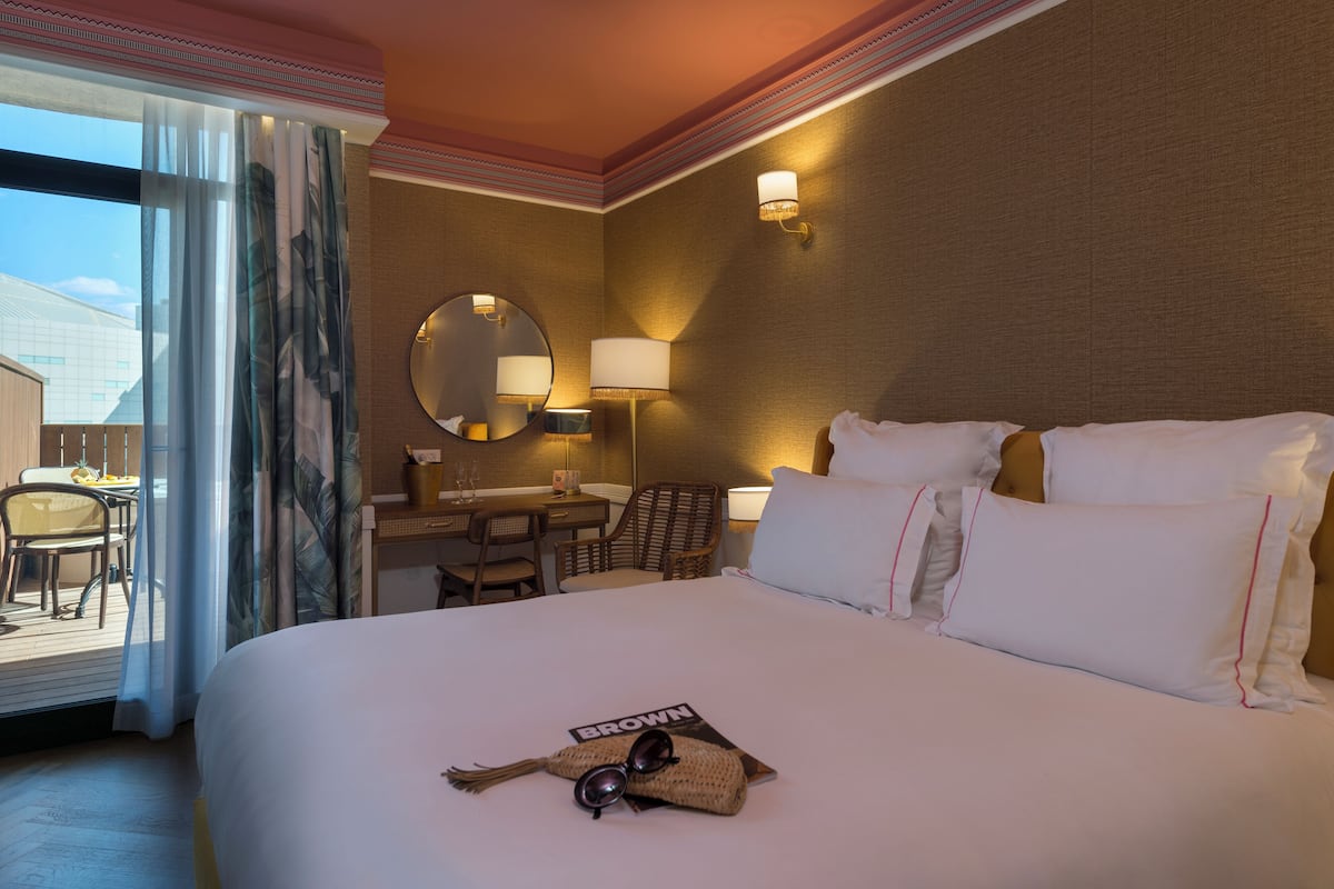 A hotel room with a large bed, a desk with a chair, a round mirror, and a view of the outside.