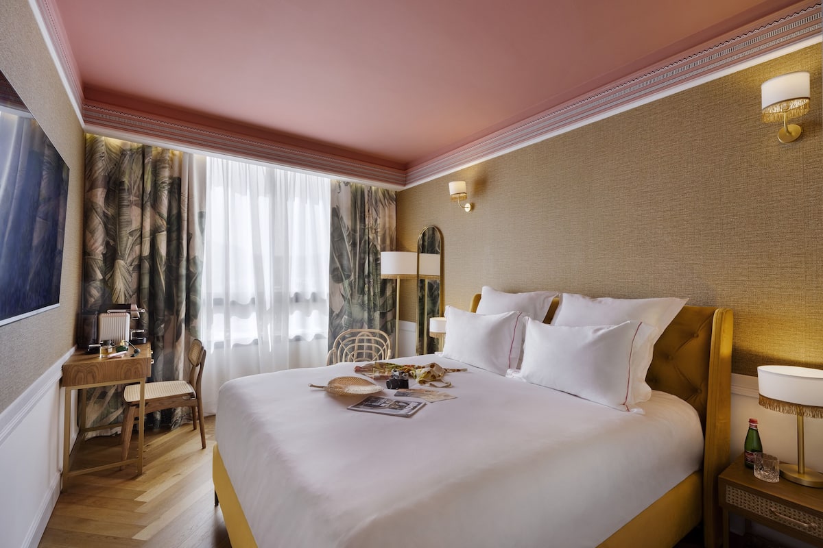 A hotel room with a large bed, a desk, a chair, and a nightstand with a lamp.