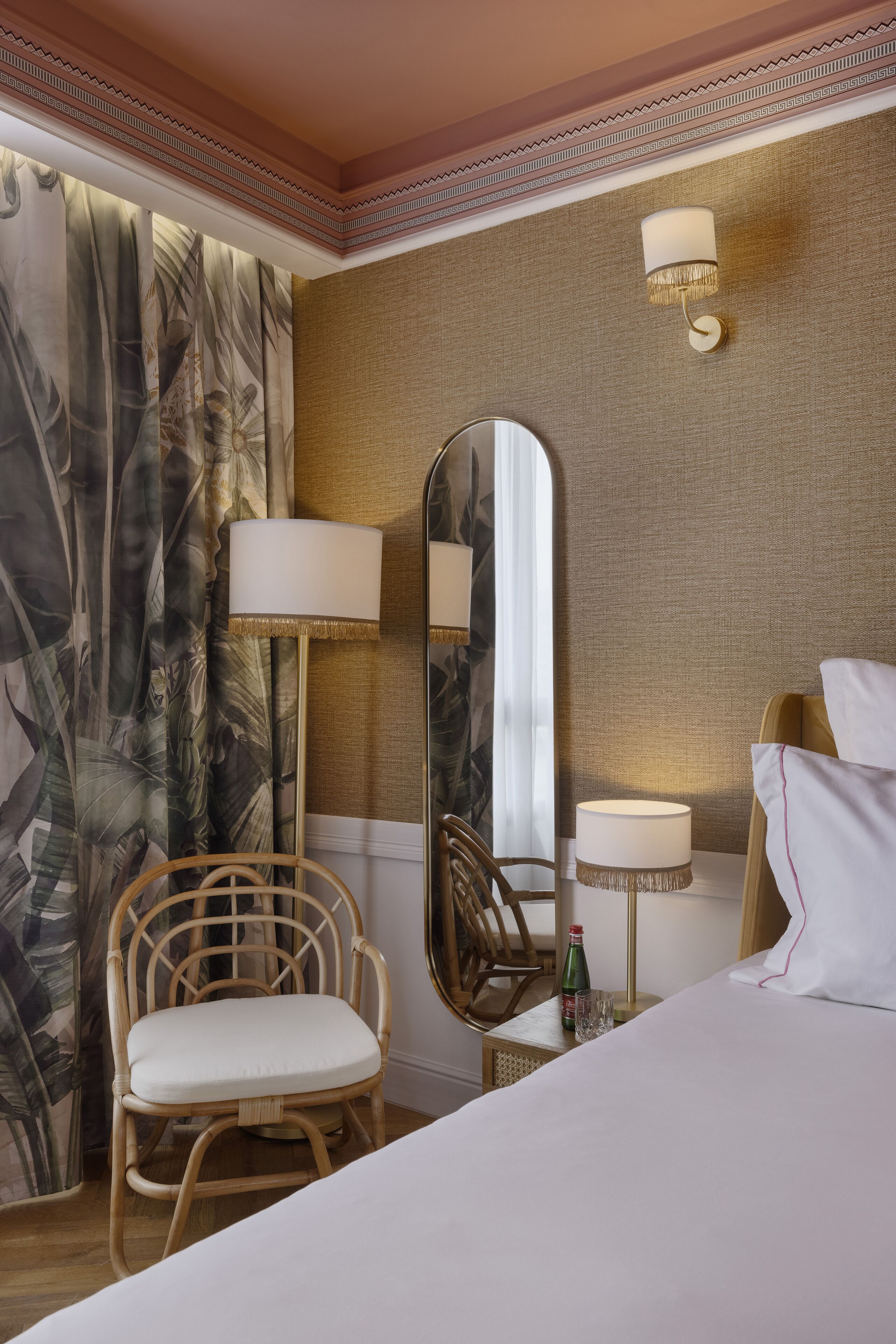Una habitación de hotel con cama, silla, espejo, lámpara y cortina estampada con hojas.