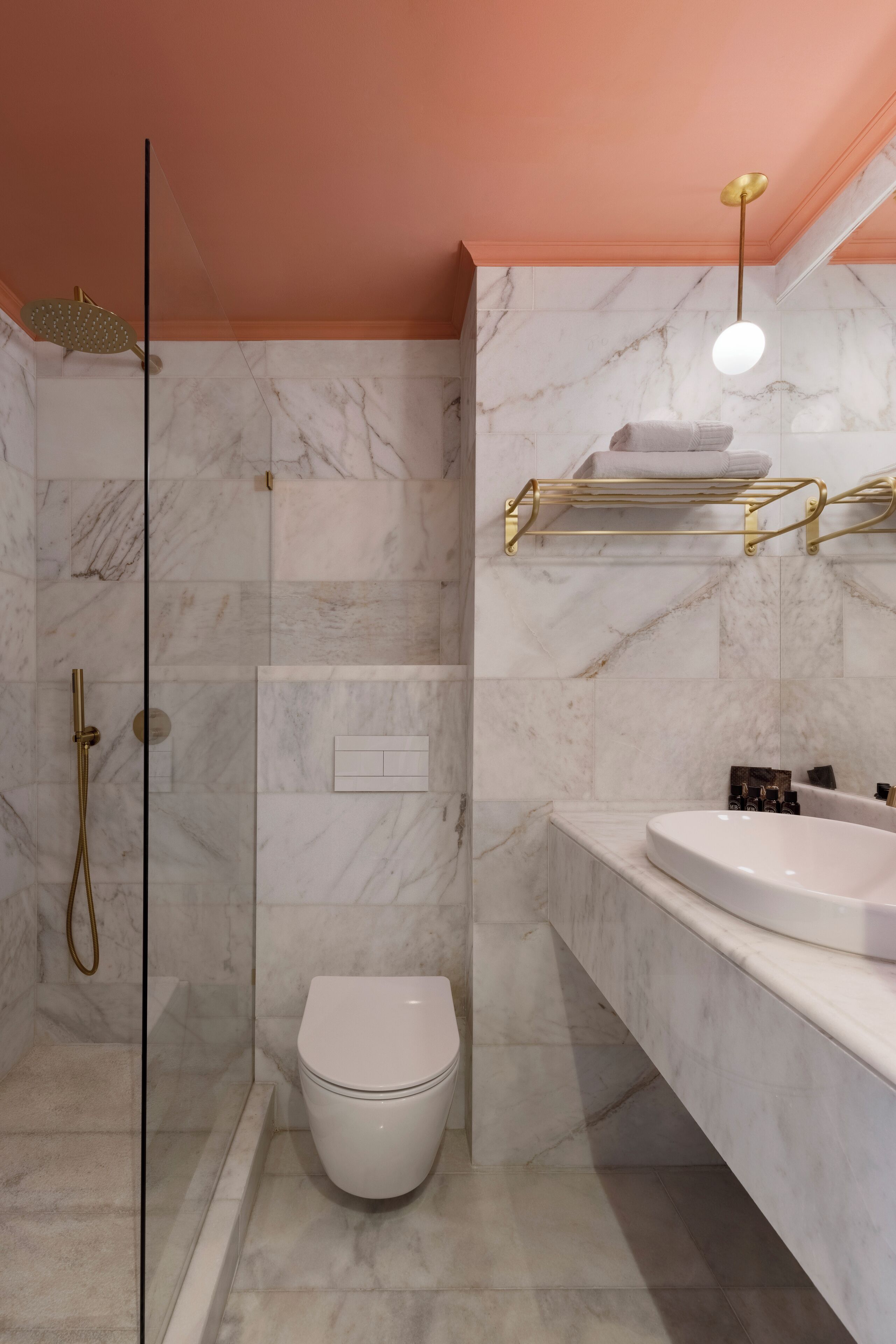 Een moderne badkamer met een marmeren vloer, een wit toilet, een douche met glazen wand en een wastafel met gouden lijst.