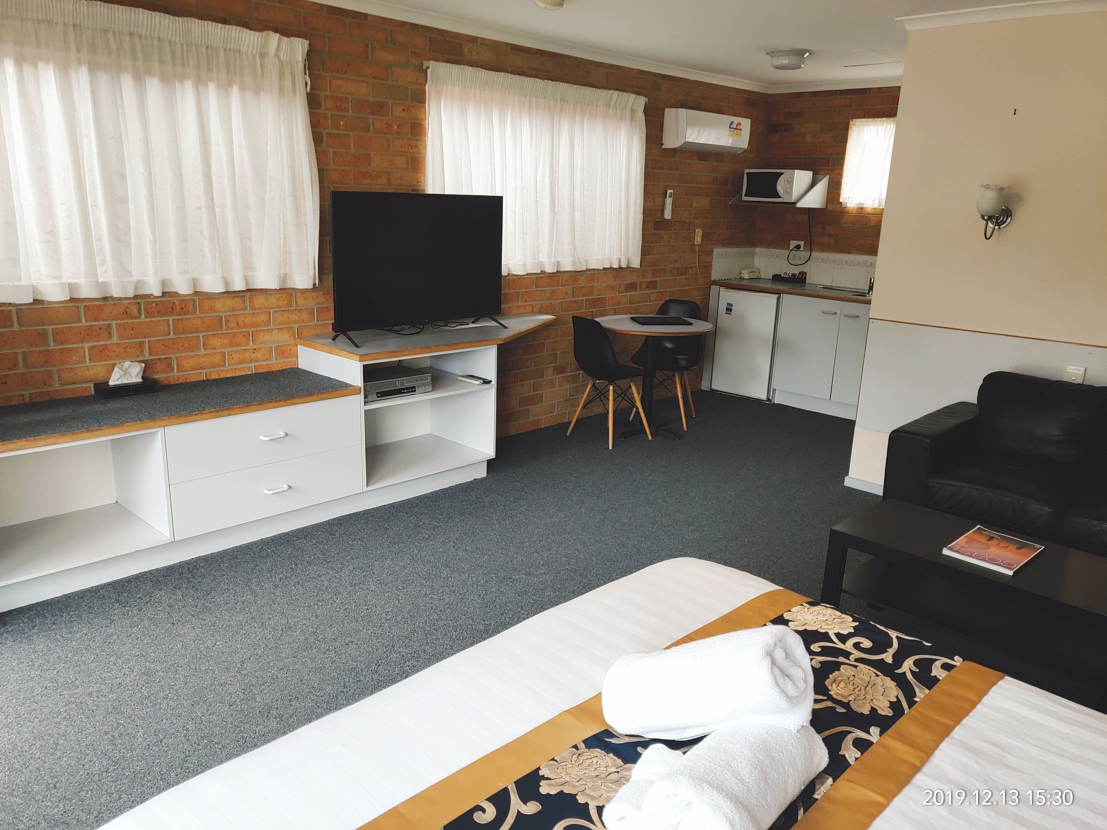 A hotel room with a bed, a TV, a desk with a chair, a kitchenette, and a black leather sofa.