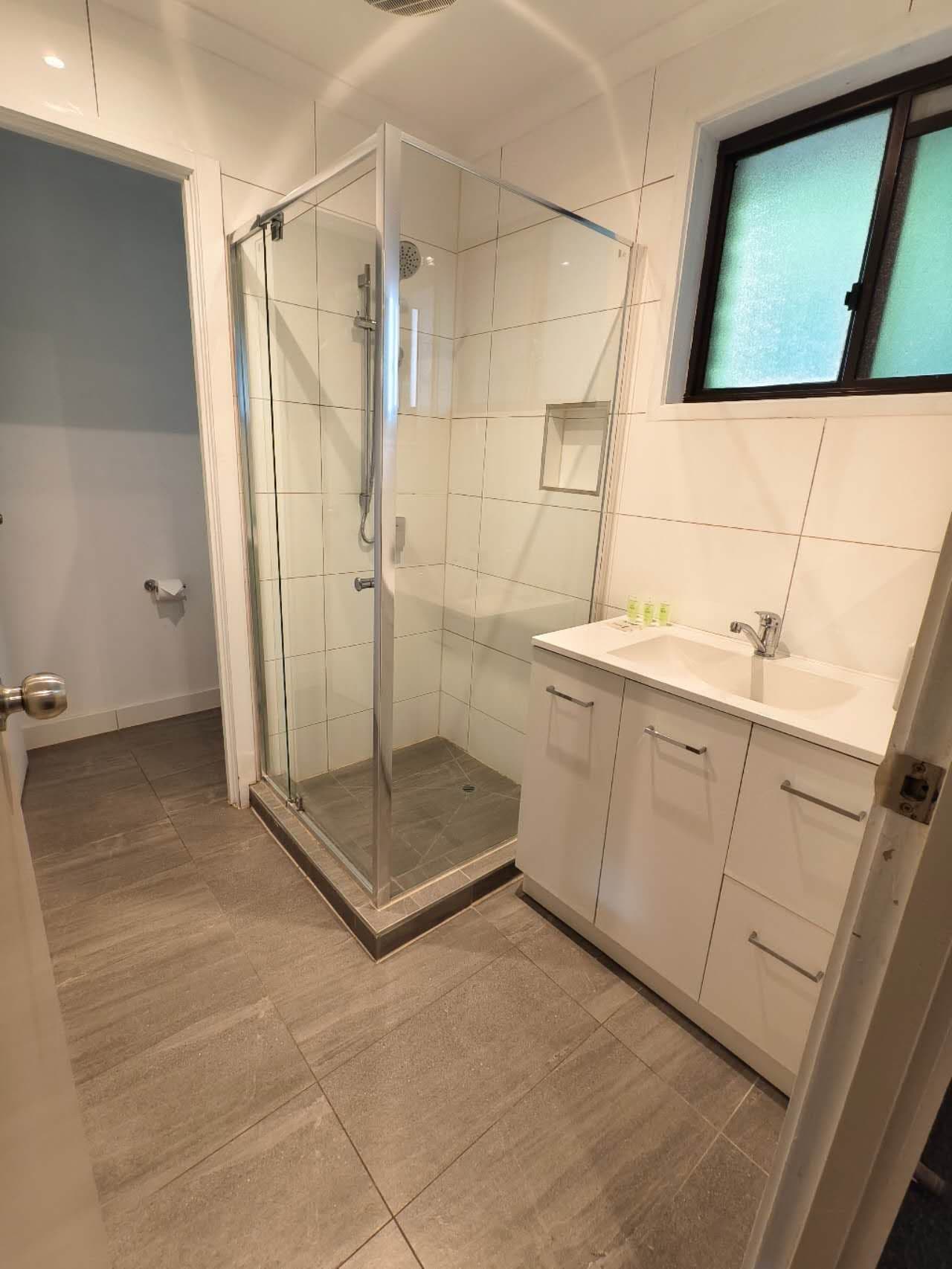 A modern bathroom with a glass shower enclosure, a white sink, and a window above the mirror.
