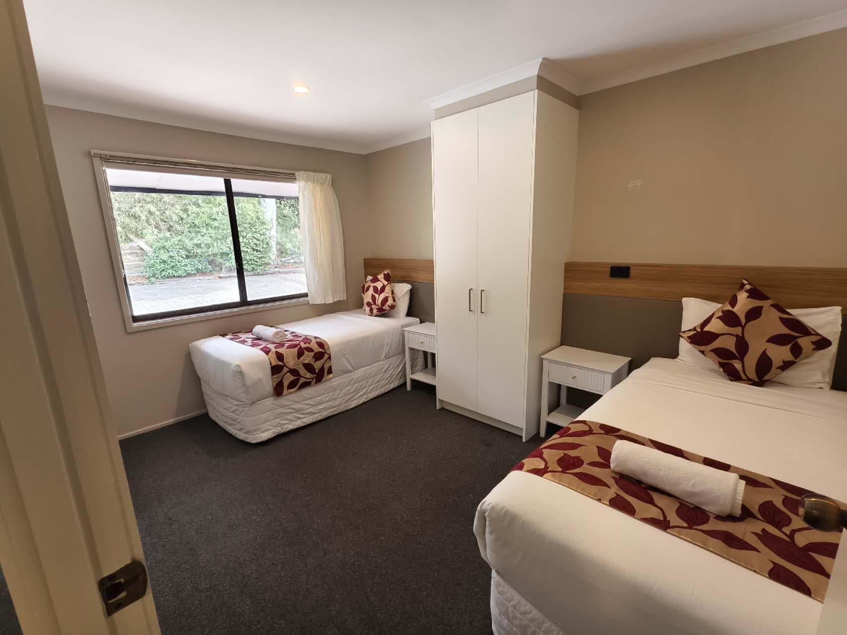 A hotel room with two beds, a wardrobe, a bedside table, and a window with a view of greenery.