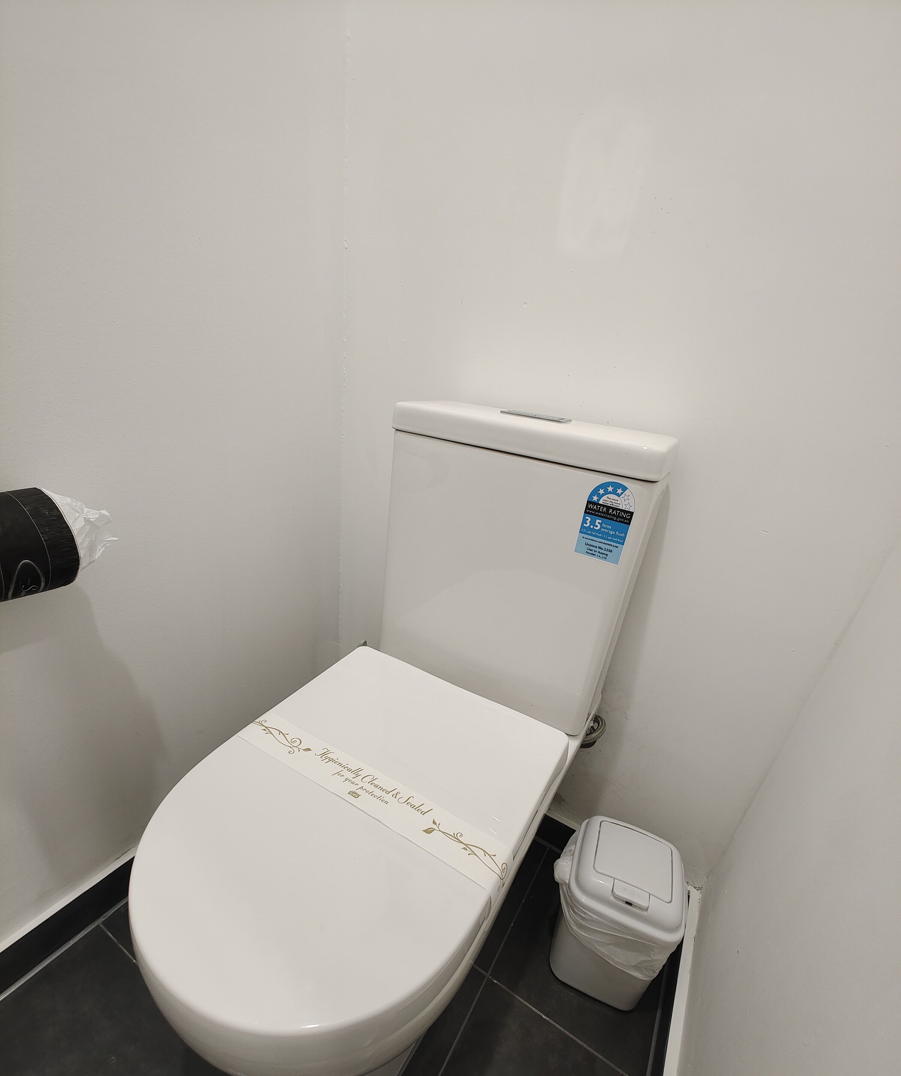 A white toilet with a water-saving sticker, a toilet paper holder, and a trash bin in a bathroom with white walls and black tiles.